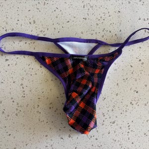 May include: A thong featuring a purple, orange, and black plaid pattern with a purple waistband and trim. A small black rectangular tag with the white text "i.defy.mishap" is centered on the front of the garment, which has a white interior lining.
