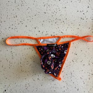 May include: A thong featuring a purple fabric base with a Halloween-themed pattern of white ghosts, orange pumpkins, and the letter R. The garment is trimmed with bright orange elastic straps and includes a black label reading defa'nSH(o)w.