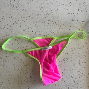 May include: A neon pink thong with bright lime green trim and thin side straps. The garment features a small white rectangular tag at the centre front with the text "i,defu'nish(a)n" printed in black, and a small size S tag on the inner fabric.