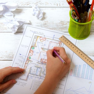 May include: A person's hands are drawing on a floor plan with a pencil. A wooden ruler and a green metal pencil holder with colored pencils are on the white wooden surface. Crumpled paper is also on the surface.