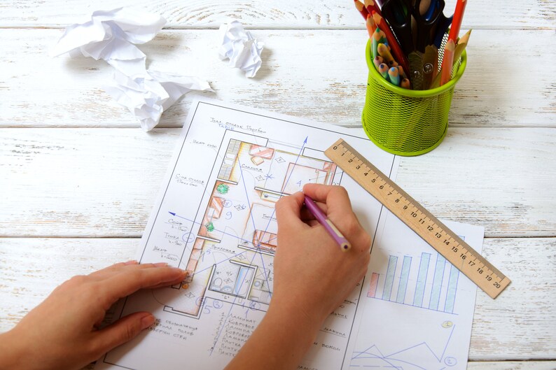 May include: A person's hands are drawing on a floor plan with a pencil. A wooden ruler and a green metal pencil holder with colored pencils are on the white wooden surface. Crumpled paper is also on the surface.