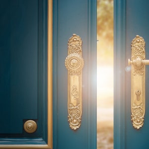 May include: A teal door with gold trim and ornate gold door hardware. The door is slightly ajar, revealing a bright, sunlit space. The hardware includes a long, decorative plate with a round knob and a handle with a keyhole.