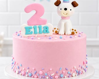 May include: A round pink frosted cake decorated with a white dog figurine wearing a blue and purple striped party hat, a pink number two, and a white bone-shaped plaque with the name Ella in blue letters, finished with colorful sprinkles.