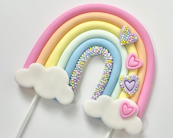 May include: A pastel rainbow cake topper with a white cloud on each end and purple hearts. The rainbow is decorated with pastel sprinkles.