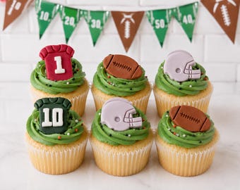 May include: Six cupcakes with green frosting and small sprinkles, topped with edible fondant decorations including red and dark green jerseys with numbers 1 and 10, brown footballs, and light gray football helmets, arranged in two rows.