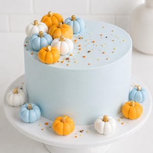 May include: A cylindrical cake with smooth light blue icing, decorated with small orange, white, and light blue pumpkins featuring gold stems. The top and base are accented with scattered gold sprinkles, presented on a white cake stand.