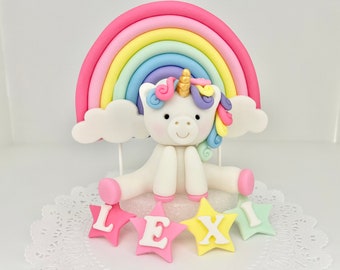 May include: A white unicorn cake topper with a rainbow, clouds, and the name "LEXI" in pink, yellow, and green stars.