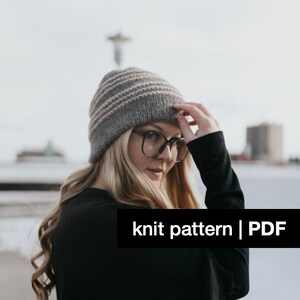 May include: A gray and beige knit beanie with horizontal stripes. The image also includes the text "knit pattern | PDF". The person in the image is wearing glasses and a black long-sleeve shirt.