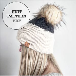 May include: A knitted beanie hat with a cream-coloured base, a dark grey band, and a large faux fur pom-pom. The hat has a small leather tag with a mountain design. Text on the image reads "KNIT PATTERN PDF".