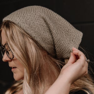 May include: A woman wearing a light brown knitted beanie hat. The hat is slouchy and has a simple design.