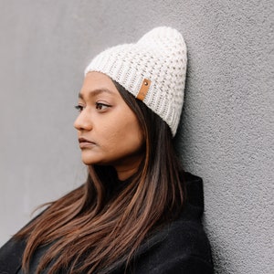 May include: Off-white knitted beanie hat with a textured design and a small brown leather tag. The hat is worn by a person with long brown hair and a black coat, leaning against a grey wall. A stylish winter accessory.