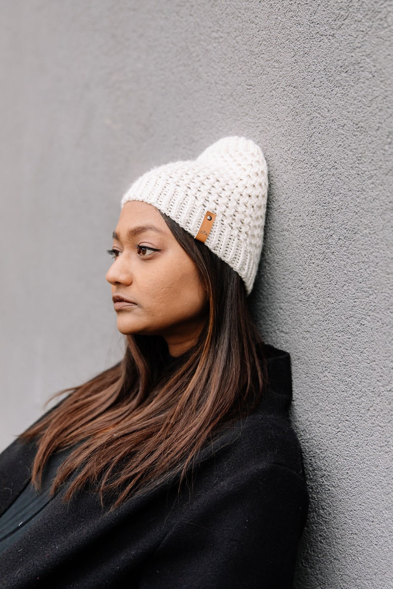 May include: Off-white knitted beanie hat with a textured design and a small brown leather tag. The hat is worn by a person with long brown hair and a black coat, leaning against a grey wall. A stylish winter accessory.