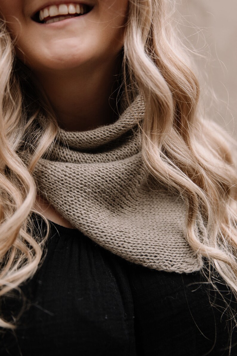 May include: A close-up of a woman wearing a brown knitted scarf. The scarf is folded in a triangular shape and is worn around the neck.