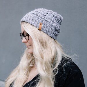 May include: A light grey knitted beanie hat with a brown leather tag. The hat is made of a thick, textured yarn and has a ribbed brim. The beanie is worn by a person with long blonde hair and black glasses.