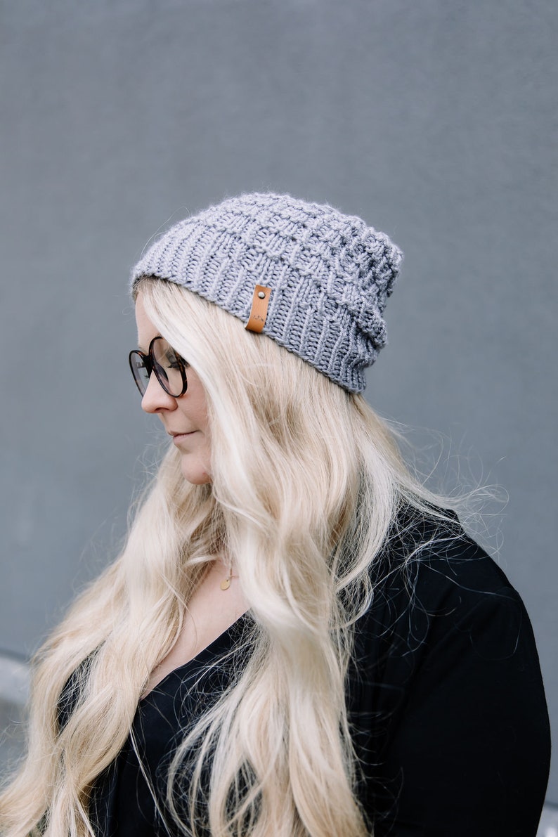 May include: A light grey knitted beanie hat with a brown leather tag. The hat is made of a thick, textured yarn and has a ribbed brim. The beanie is worn by a person with long blonde hair and black glasses.