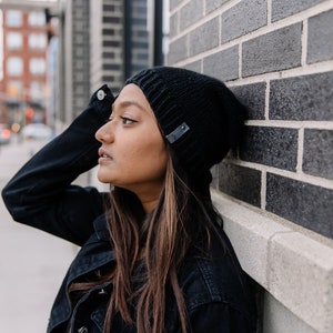 May include: Black knitted beanie with a small leather tag detail. The beanie is worn with a black denim jacket. The background features a brick wall and a city street.