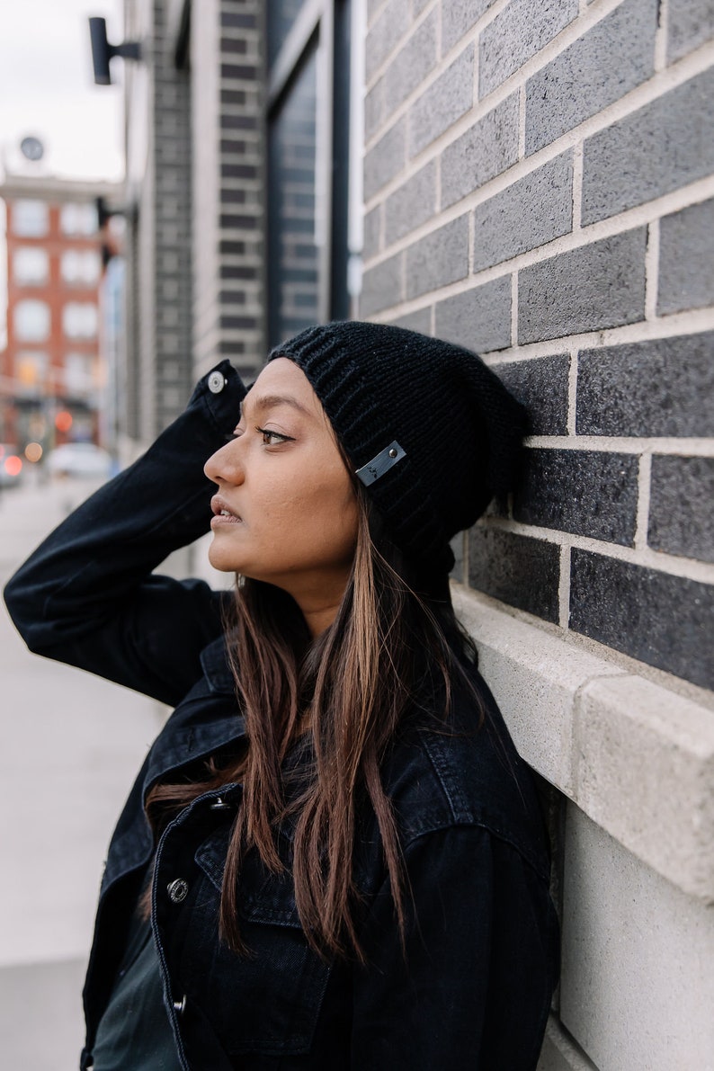 May include: Black knitted beanie with a small leather tag detail. The beanie is worn with a black denim jacket. The background features a brick wall and a city street.