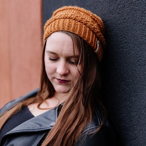 May include: A mustard-coloured knitted beanie hat with a ribbed design. The hat is worn by a person with long, auburn hair and a black leather jacket. The background features a dark grey wall and a reddish-brown wall.
