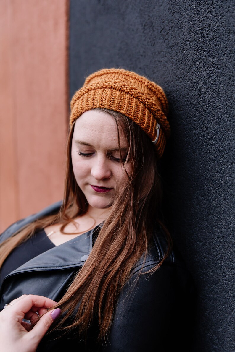 May include: A mustard-coloured knitted beanie hat with a ribbed design. The hat is worn by a person with long, auburn hair and a black leather jacket. The background features a dark grey wall and a reddish-brown wall.