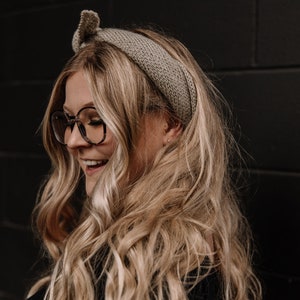 May include: A woman with long, wavy blonde hair is wearing a light brown knitted headband and round, tortoiseshell glasses. She is smiling and looking down.