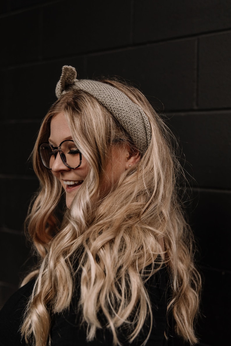 May include: A woman with long, wavy blonde hair is wearing a light brown knitted headband and round, tortoiseshell glasses. She is smiling and looking down.