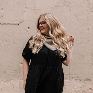 May include: A woman with long blonde hair wearing a black tiered dress and a light brown bandana. She is wearing blue-rimmed glasses and smiling.