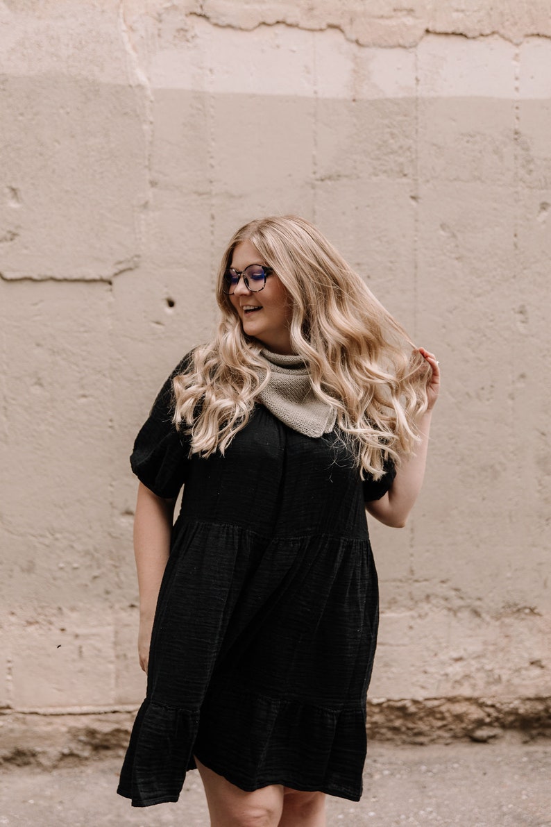 May include: A woman with long blonde hair wearing a black tiered dress and a light brown bandana. She is wearing blue-rimmed glasses and smiling.