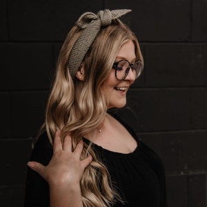 May include: A woman with long blonde hair wearing a brown knitted headband with a bow and black framed glasses. She is wearing a black top.