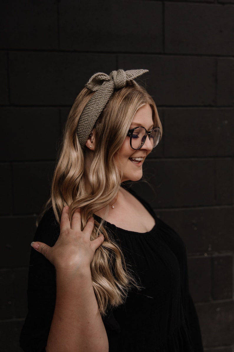 May include: A woman with long blonde hair wearing a brown knitted headband with a bow and black framed glasses. She is wearing a black top.