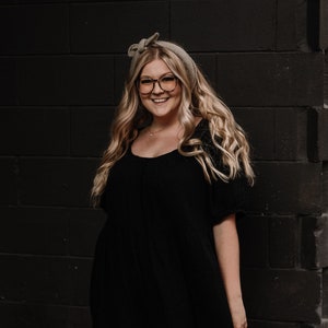 May include: A woman with long blonde hair wearing a black dress and glasses, standing in front of a dark brick wall.