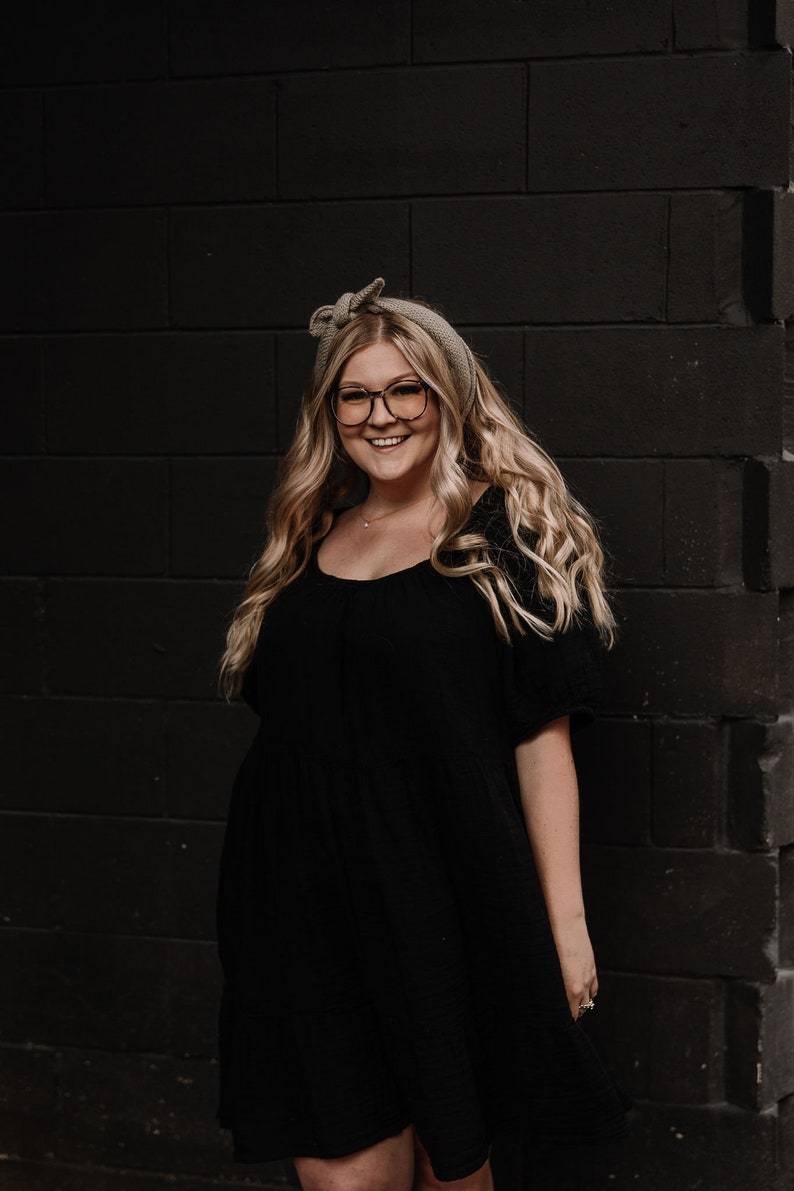 May include: A woman with long blonde hair wearing a black dress and glasses, standing in front of a dark brick wall.