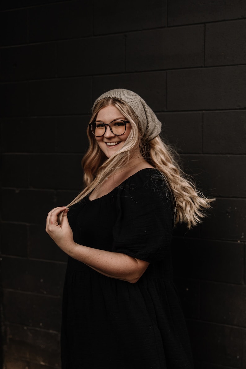 May include: A woman with long blonde hair wearing a grey beanie and black dress, smiling at the camera.