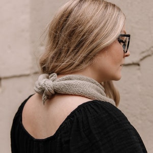 May include: A woman wearing a black top with a light brown scarf tied around her neck. She is wearing black framed glasses and has long blonde hair.