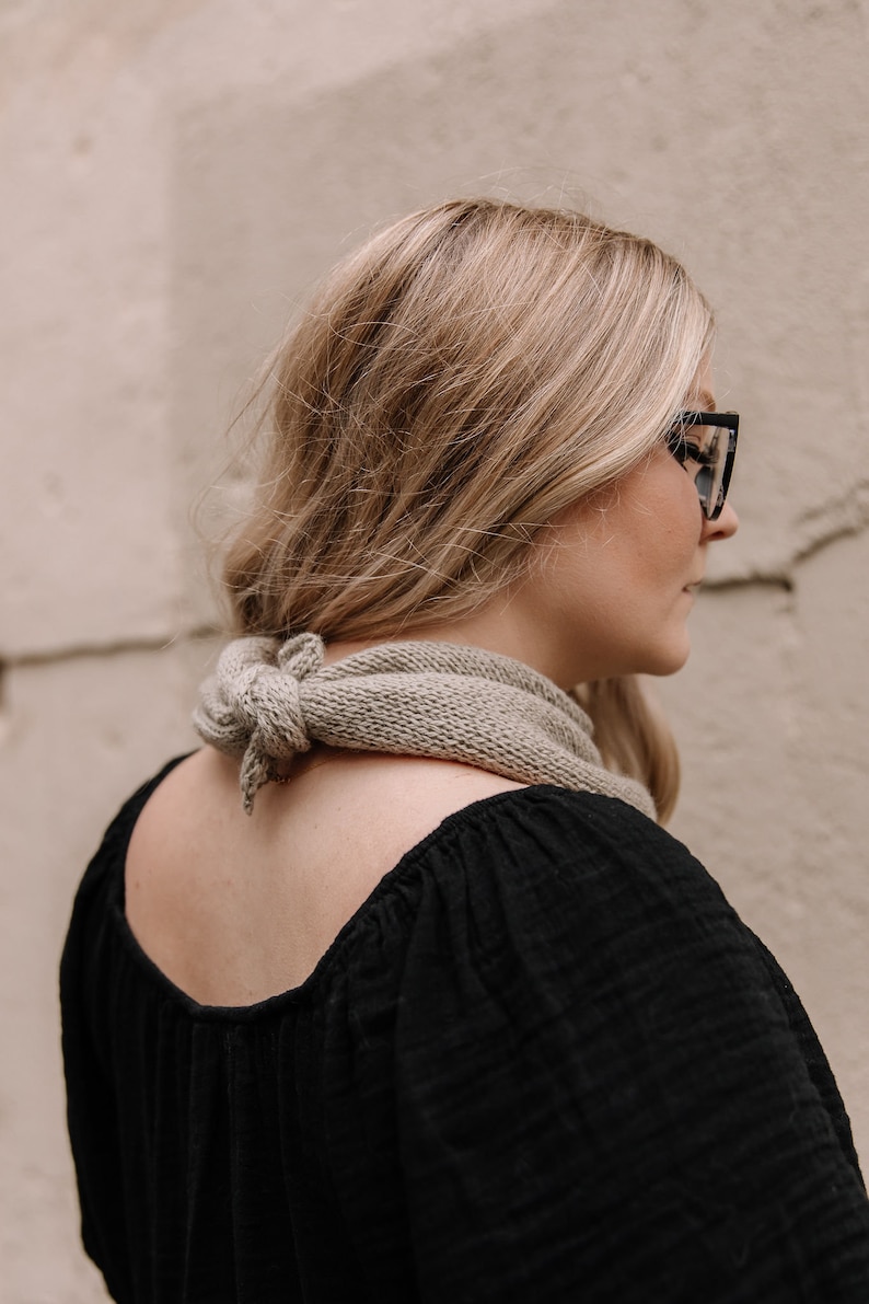 May include: A woman wearing a black top with a light brown scarf tied around her neck. She is wearing black framed glasses and has long blonde hair.