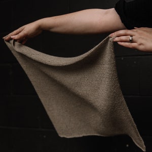 May include: A hand holding a light brown knitted scarf. The scarf is made of a soft, textured yarn and has a simple, elegant design.