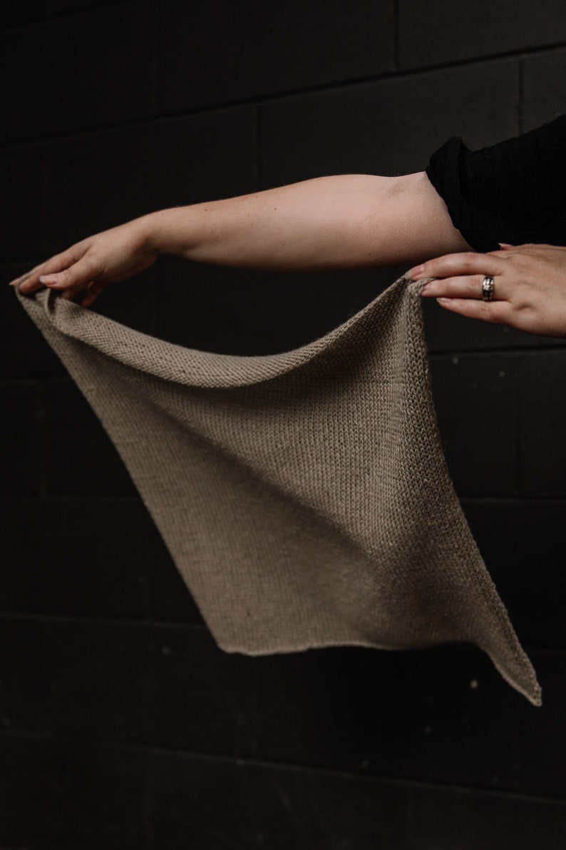 May include: A hand holding a light brown knitted scarf. The scarf is made of a soft, textured yarn and has a simple, elegant design.
