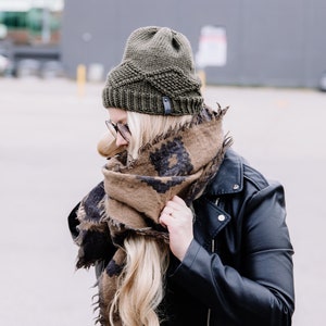 May include: An olive green knitted beanie with a leather tag is worn with a brown and black patterned scarf and a black leather jacket. The scarf is wrapped around the neck and shoulders.