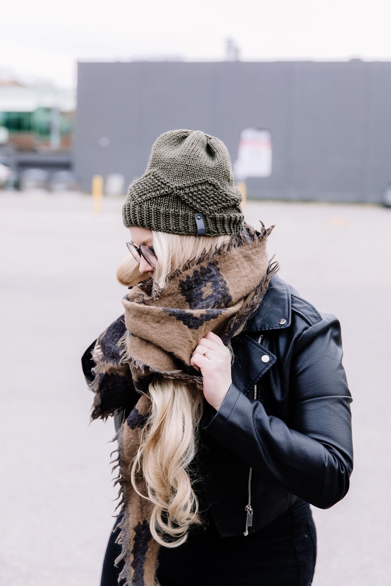 May include: An olive green knitted beanie with a leather tag is worn with a brown and black patterned scarf and a black leather jacket. The scarf is wrapped around the neck and shoulders.