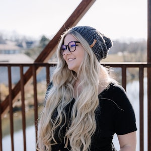 May include: A woman wearing a grey knitted beanie, black-framed glasses, and a black top. She is smiling and looking to the side. The background includes a bridge and a sunny sky.