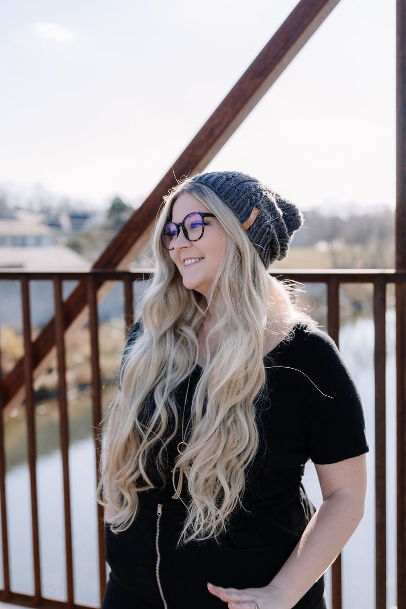 May include: A woman wearing a grey knitted beanie, black-framed glasses, and a black top. She is smiling and looking to the side. The background includes a bridge and a sunny sky.