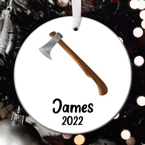 May include: White round Christmas ornament with a cartoon image of an axe and the text "James 2022".