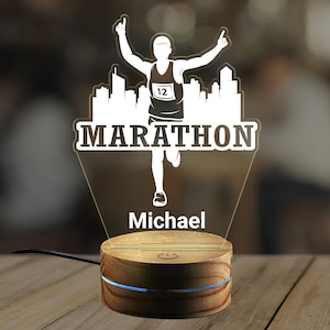 Op de afbeelding: Een wit silhouet van een hardloper met het woord "MARATHON" en het nummer "12" op zijn borst. De hardloper rent door een stadsbeeld. De afbeelding staat op een houten voet met de naam "Michael" eronder.