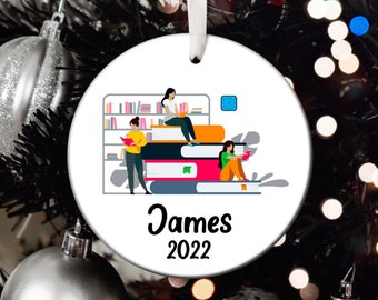 May include: White round Christmas ornament with a colorful illustration of three people sitting on a stack of books. The ornament has the text "James 2022" printed on it.