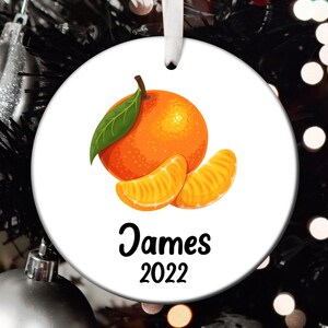 May include: A white round Christmas ornament with a picture of a whole orange and two orange slices. The ornament has the name "James" and the year "2022" printed on it.
