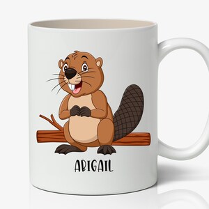 May include: White ceramic mug featuring a cartoon beaver with brown fur, a large tail, and a happy expression. The beaver is sitting on a log, and the name "ABIGAIL" is printed below. The mug has a white handle.