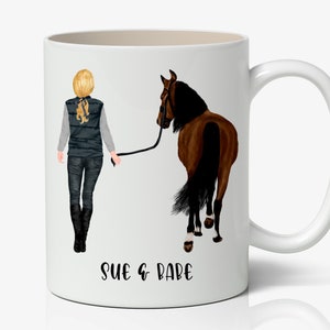 May include: White ceramic mug with a brown horse and a woman with blonde hair walking with the horse. The text "SUE & BABE" is printed on the bottom of the mug.