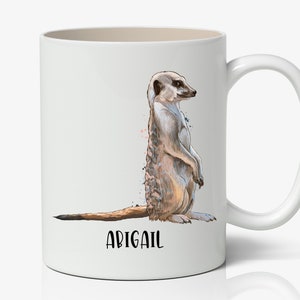 May include: White ceramic mug with a watercolor illustration of a meerkat standing on its hind legs. The mug has a black handle and the name "Abigail" is printed on the bottom.
