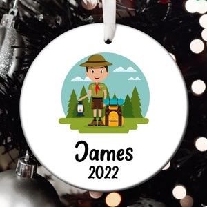 May include: A white round ornament with a cartoon illustration of a scout holding a lantern and a backpack. The ornament has the text "James 2022" printed on it.