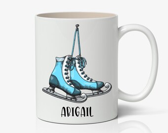 May include: White ceramic mug with a blue and white illustration of ice skates hanging from a string. The text "ABIGAIL" is printed in black at the bottom of the mug.