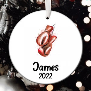 May include: White ceramic Christmas ornament with a watercolor illustration of red boxing gloves and a silver knife. The name "James" and the year "2022" are printed in black script. A white ribbon is attached for hanging.